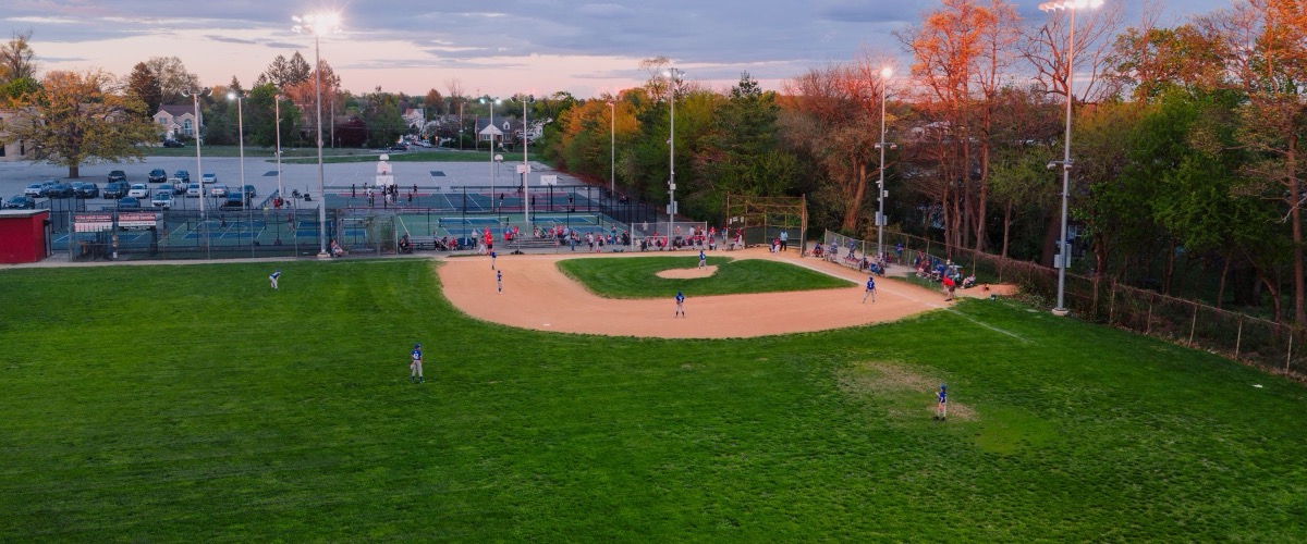 A youth ballfield under the lights at dusk, seen from above, with courts and a parking lot behind it